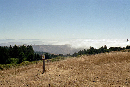 [Atop a hill with a trail post in the foreground looking out at the mountains which are partially covered with clouds.]